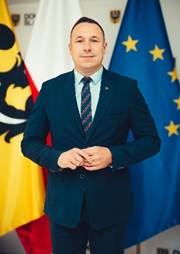 Na pierwszym planie stoi m�czyzna o neutralnym wyrazie twarzy i br�zowych, kr�tkoprzystrzy�onych w�osach. Ma na sobie granatowy garnitur i jasn� koszul�. W tle trzy flagi: Polski, Dolnego �l�ska i Unii Europejskiej.
